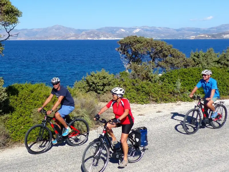 Drei Personen fahren mit Fahrrädern auf einer Küstenstraße. Im Hintergrund sind das Meer und Berge zu sehen.