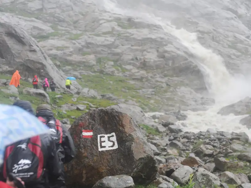 Wanderer auf dem Europäischen Fernwanderweg E5, markiert durch einen Stein mit der E5-Kennzeichnung, passieren einen Wasserfall im Regen.