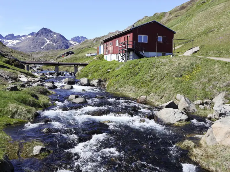 Ein Fluss fließt durch eine bergige Landschaft mit einer roten Hütte und einer Brücke im Vordergrund.