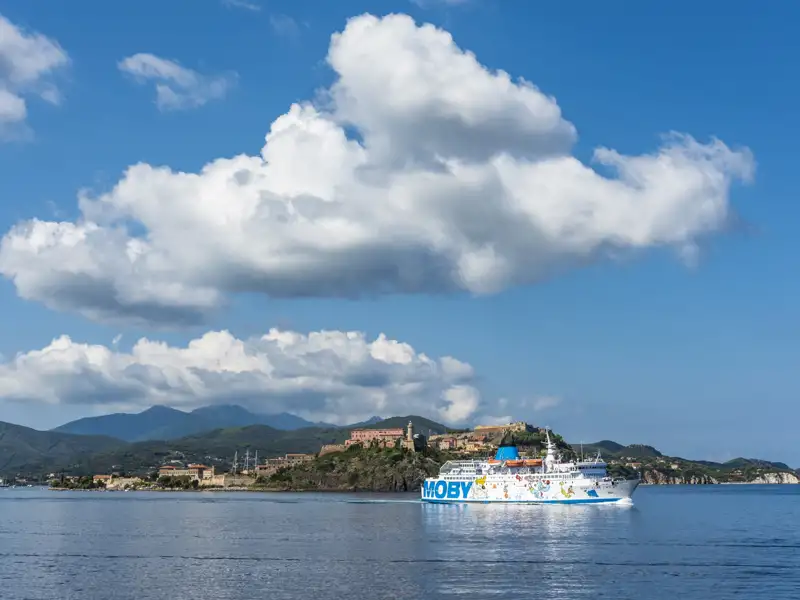 Moby-Fähre auf dem Wasser unterwegs, Inselpassage mit Gebäuden und Bergen im Hintergrund.