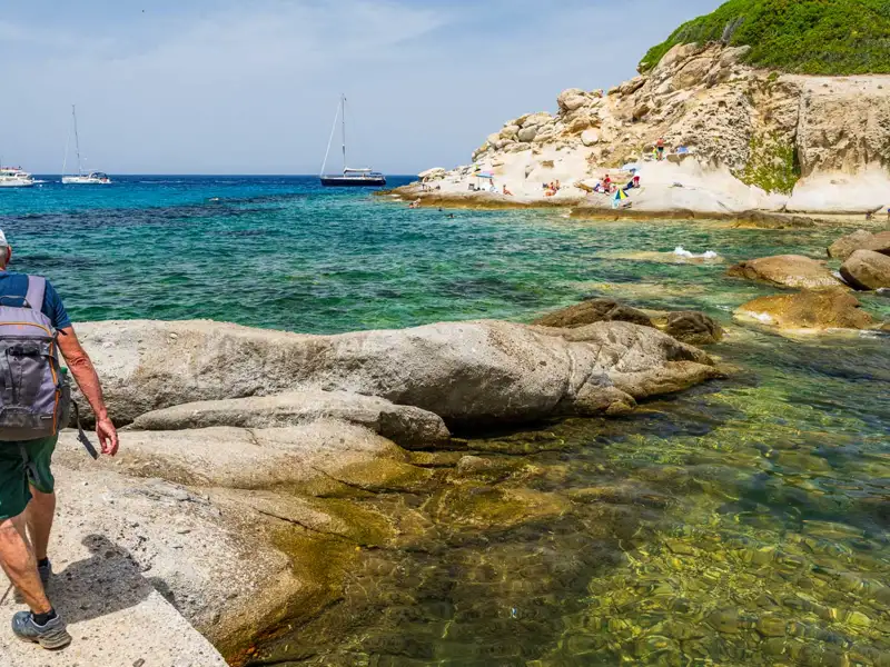 Wanderung entlang einer felsigen Küste mit türkisfarbenem Wasser und Segelbooten im Hintergrund.