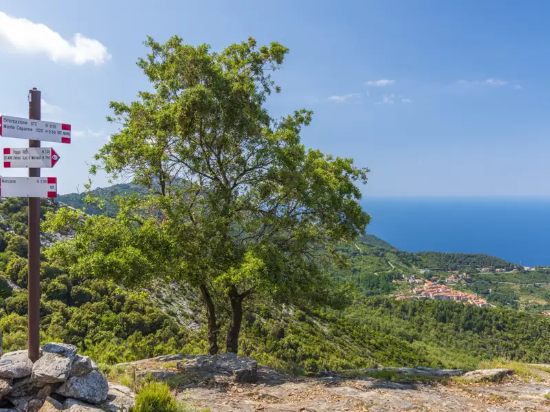 Wegweiser mit Richtungsangaben nach Marciana, Poggio und Monte Capanne auf Elba. Im Hintergrund das Meer und eine Ortschaft.
