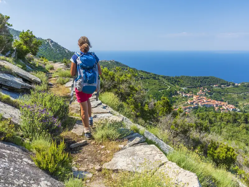 Wanderin mit Rucksack auf einem steinigen Küstenpfad. Im Hintergrund das blaue Meer und ein Dorf.