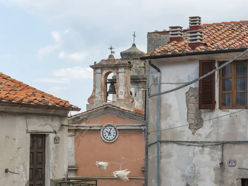 Kirchturm mit Uhr und Glocke inmitten von Häusern mit Terrakottadächern in einem italienischen Dorf.