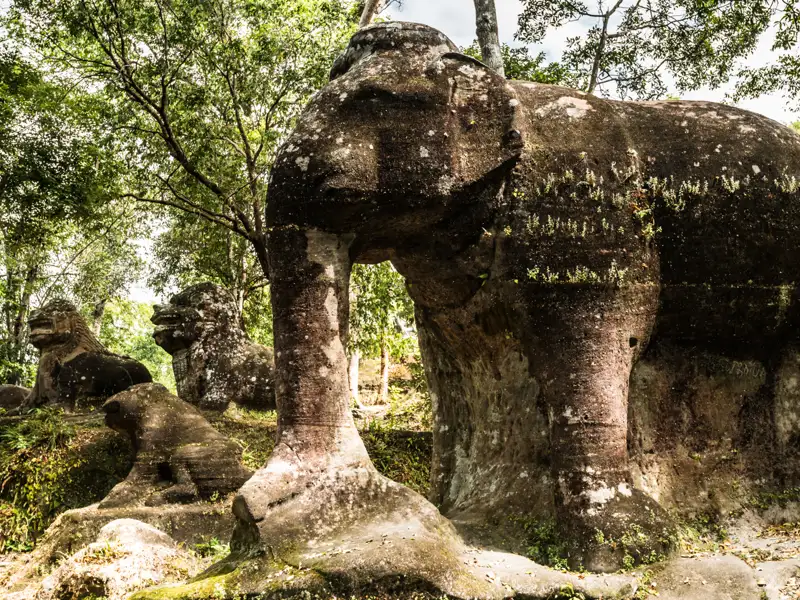 Alte steinerne Elefanten- und Löwenstatuen, umgeben von üppiger Vegetation.
