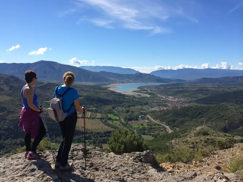 Zwei Wanderer mit Wanderstöcken pausieren auf einem Felsvorsprung und genießen den Ausblick auf das Tal, den See und das Dorf in der Ferne.