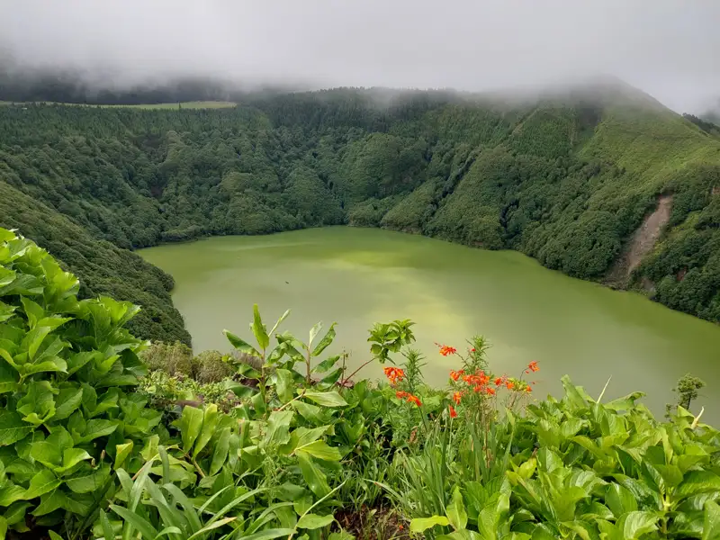 Panoramablick auf einen grünen Kratersee, umgeben von dichter Vegetation und nebligen Hügeln.