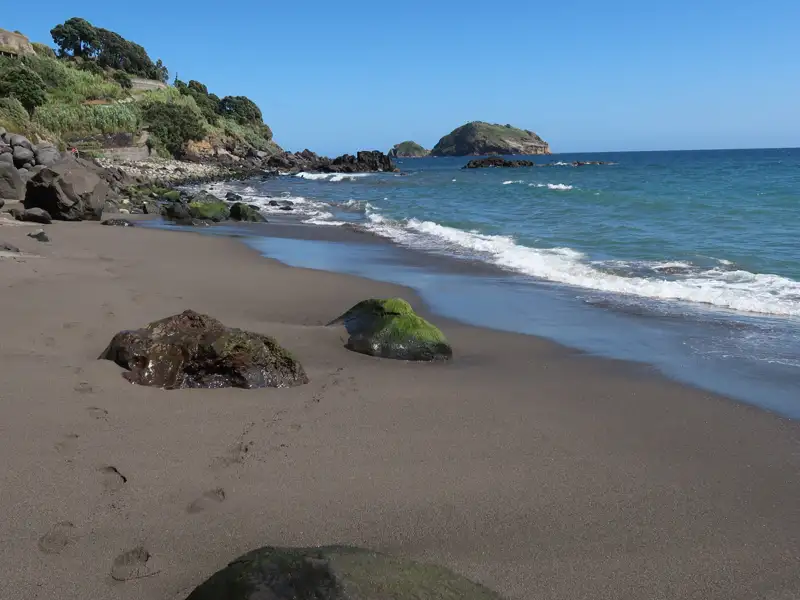 Ansicht eines dunklen Sandstrandes mit Felsen, bewachsener Küste und einer kleinen Insel im Hintergrund.