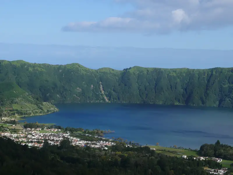 Ausblick auf einen See, umgeben von bewaldeten Hügeln. Eine kleine Ortschaft ist am Ufer sichtbar.