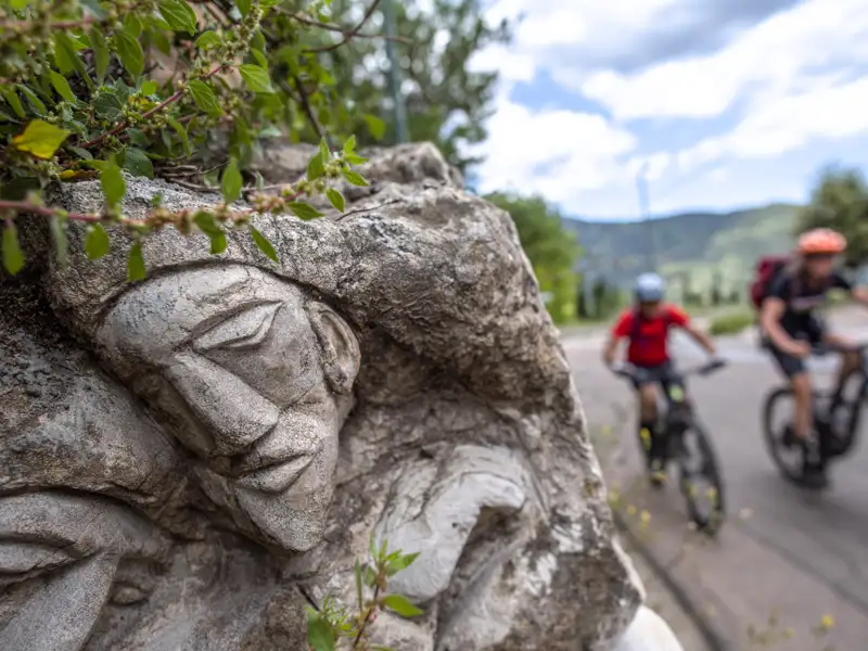 Nahaufnahme einer Steinskulptur, während Mountainbiker auf einem Weg im Hintergrund fahren.
