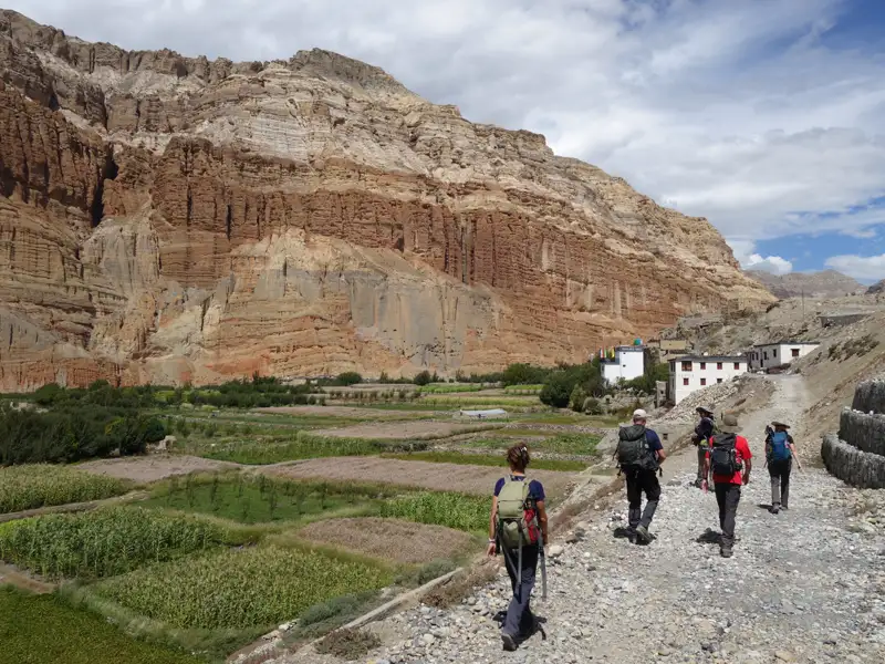 Wandergruppe auf einem Bergpfad mit Blick auf terrassierte Felder und ein Gebäude in der Ferne. Die Felswand im Hintergrund verdeutlicht die landschaftliche Schönheit der Wanderung.