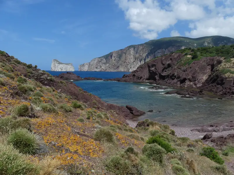 Sicht auf eine felsige Küstenlandschaft mit rötlichen Felsen und spärlicher Vegetation. Im Hintergrund sind zwei markante Felseninseln im Meer sichtbar.