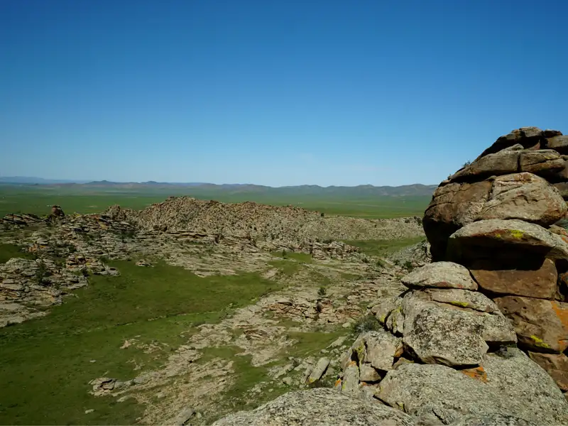 Felsige Landschaft mit grünen Wiesen und Hügeln im Hintergrund unter blauem Himmel.