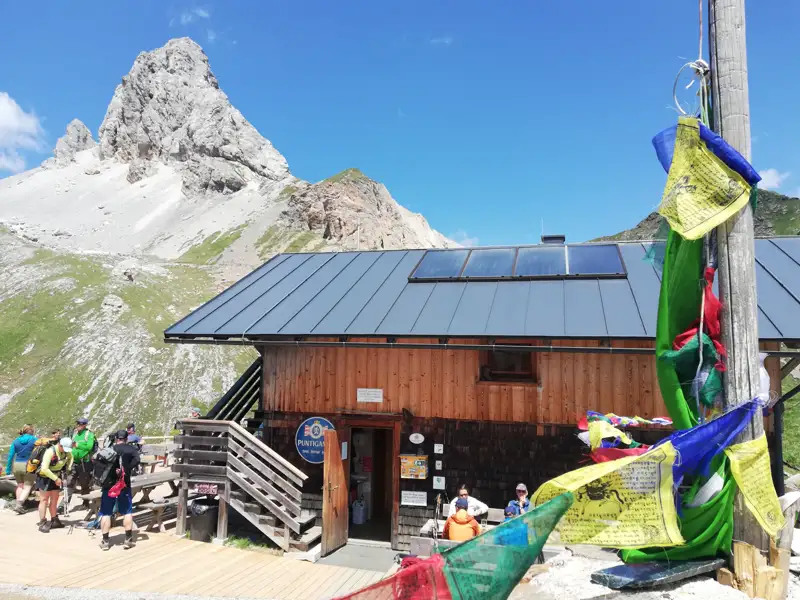 Berghütte in den Bergen mit Solarpanelen und Gebetsfahnen, im Hintergrund ein Berg.