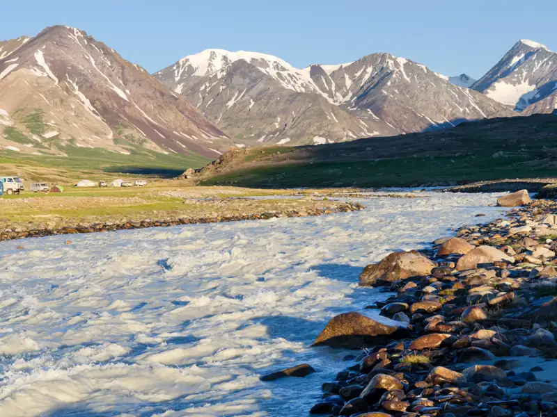 Schneebedeckte Berge, ein schnell fließender Fluss und ein Basislager im Vordergrund.