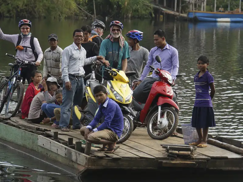 Reisende und Einheimische überqueren einen Fluss auf einer Fähre. Fahrräder und Motorroller werden transportiert.