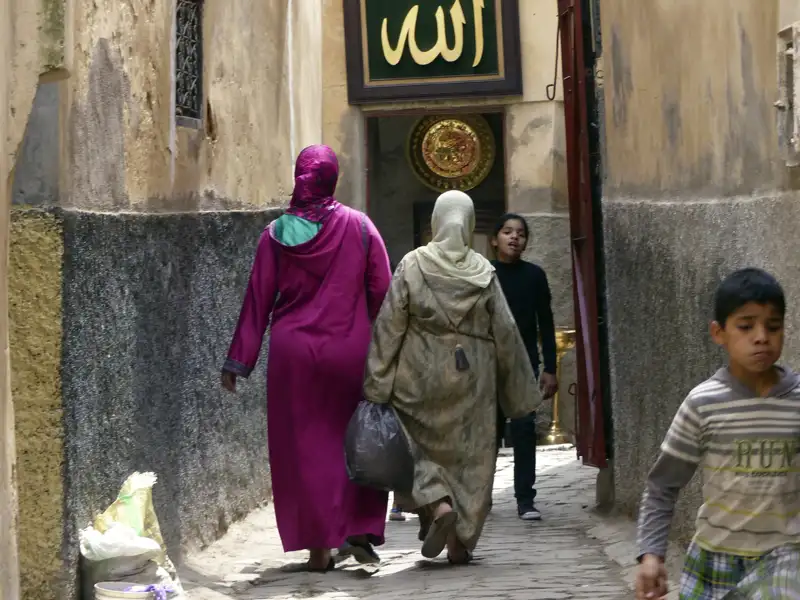 Zwei Frauen in traditionellen Gewändern gehen durch eine enge Gasse. Im Hintergrund sind Kinder und ein verzierter Türrahmen zu sehen.