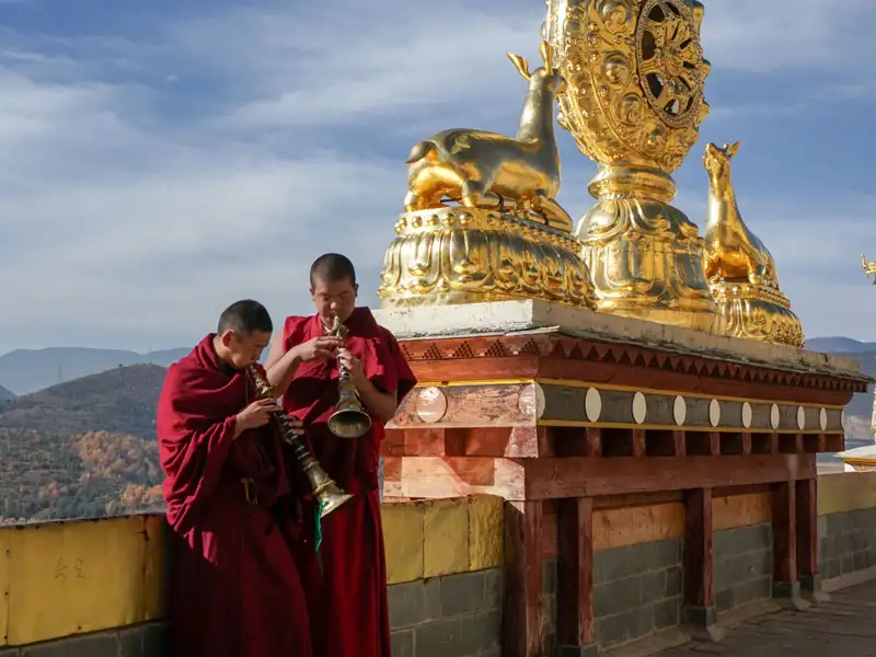 Zwei Mönche spielen traditionelle Instrumente vor einer goldenen Statue mit Dharma-Rad und Hirschen, die einen wichtigen Aspekt des buddhistischen Glaubens symbolisieren.