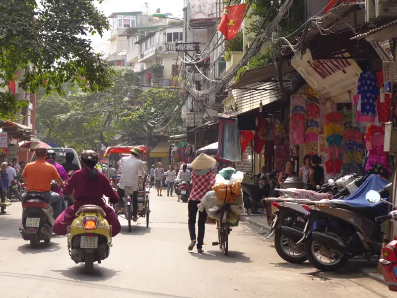 Straßenszene in Hanoi mit Fußgängern, Fahrradfahrern, Motorradfahrern und Händlern, die die geschäftige Atmosphäre der Stadt zeigen.