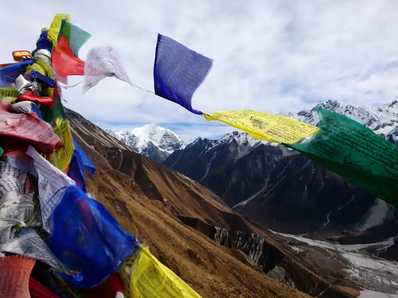Gebetsfahnen im Vordergrund, dahinter schneebedeckte Berge und ein Tal.