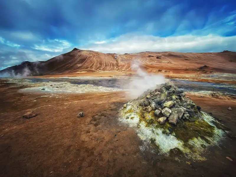 Dampfende Gesteinsformation in einer geothermalen Landschaft mit bunten Gesteinen und einem Bach im Hintergrund.