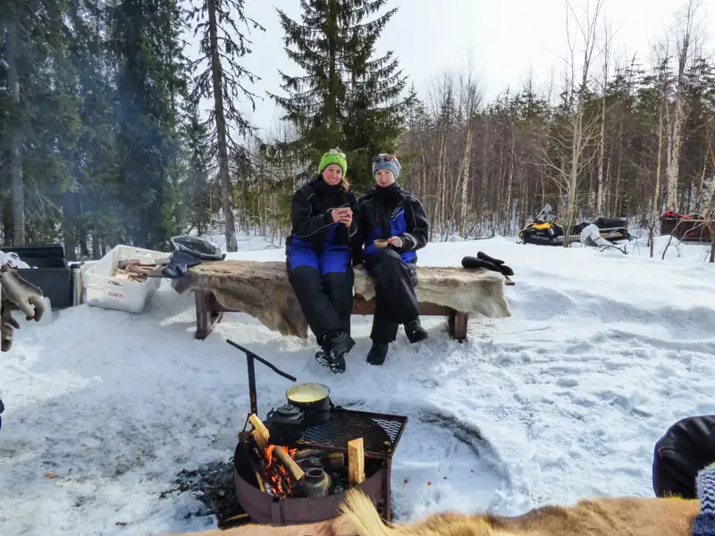 Zwei Reisende wärmen sich an einem Lagerfeuer während einer Pause von ihrer Schneemobiltour im verschneiten Wald.