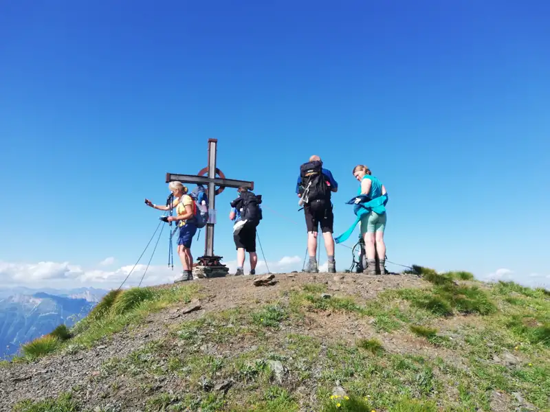 Wanderer mit Rucksäcken stehen am Gipfelkreuz und blicken in die Ferne.