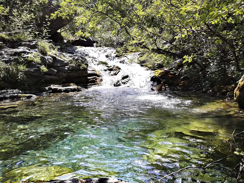 Person badet in einem klaren Flussbecken unter einem Wasserfall.