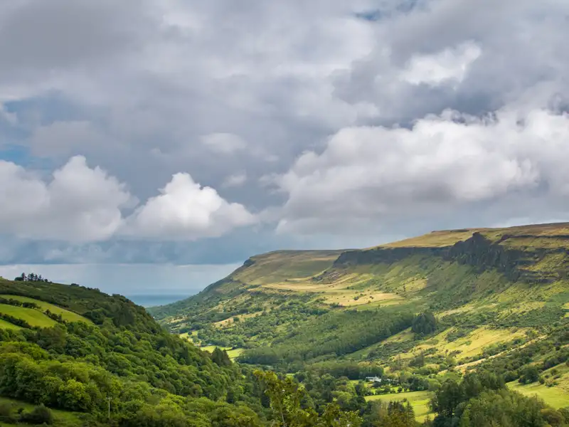 Panoramablick auf ein Tal mit üppiger Vegetation und bewaldeten Hängen unter einem bewölkten Himmel.