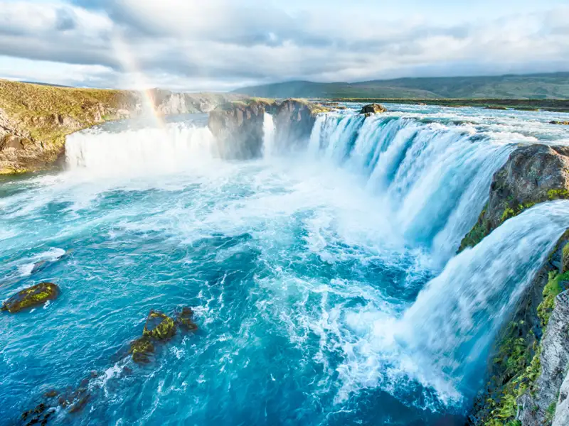 Panoramablick auf einen mehrstufigen Wasserfall mit türkisfarbenem Wasser und einem Regenbogen.