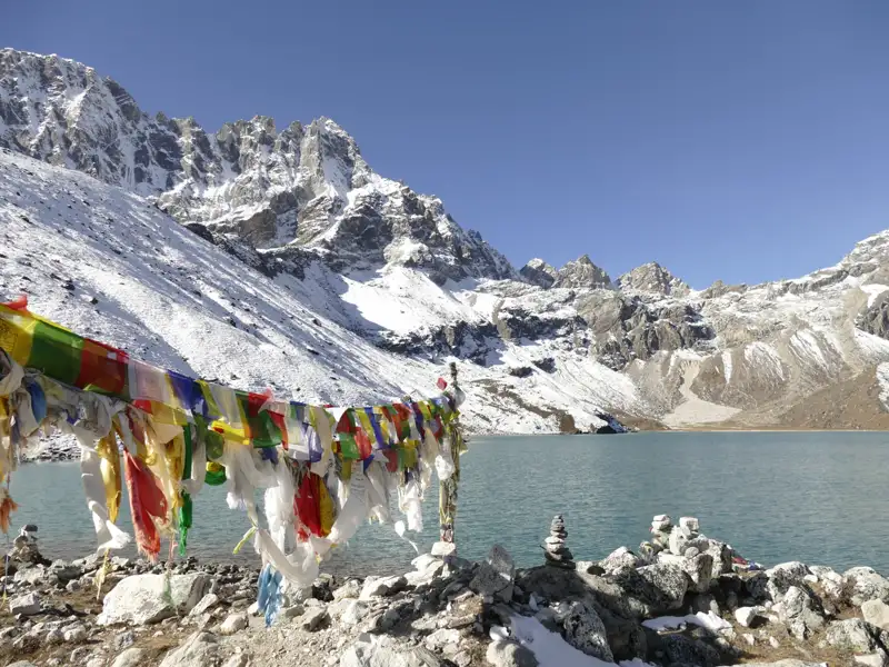Bunte Gebetsfahnen am Ufer eines Bergsees mit schneebedeckten Himalaya-Gipfeln im Hintergrund.