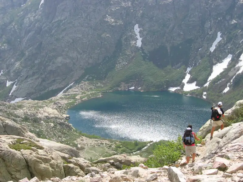 Zwei Wanderer auf einem Bergpfad. Ein Bergsee ist im Hintergrund zu sehen, umgeben von felsigen Hängen und Resten von Schneefeldern.