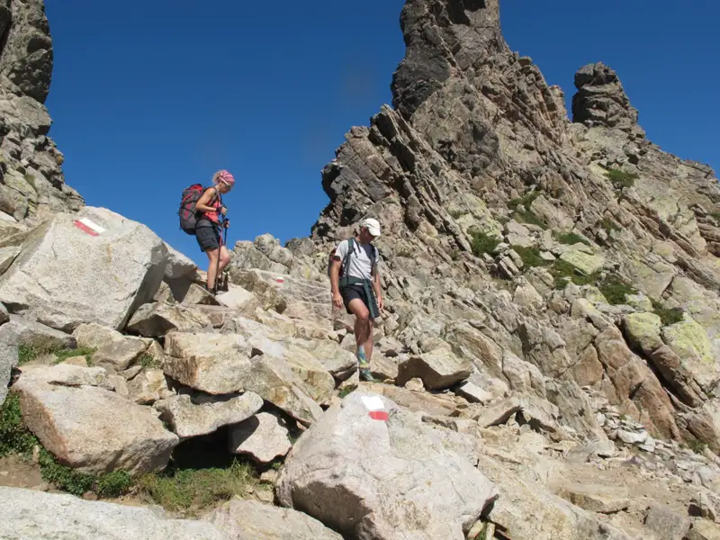 Zwei Wanderer erklimmen einen felsigen Bergpfad, markiert mit rot-weißen Wegzeichen.