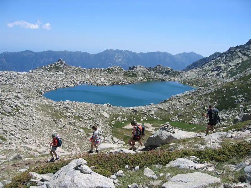 Wanderer mit Rucksäcken steigen einen felsigen Pfad hinab in Richtung eines Bergsees. Im Hintergrund erstreckt sich eine Bergkette.