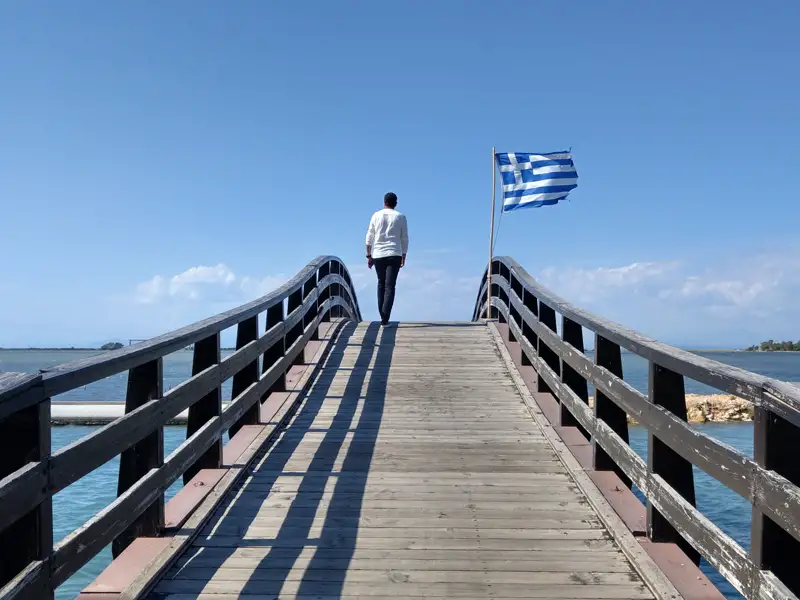 Holzbrücke mit griechischer Flagge und Blick aufs Meer.