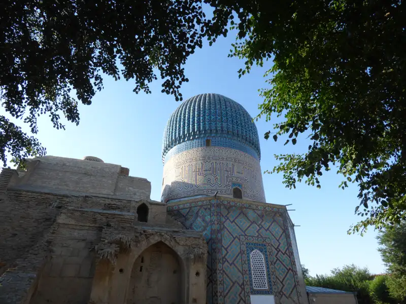 Blau-weiße Kuppel und Fassade eines Mausoleums, gesehen durch Baumzweige.