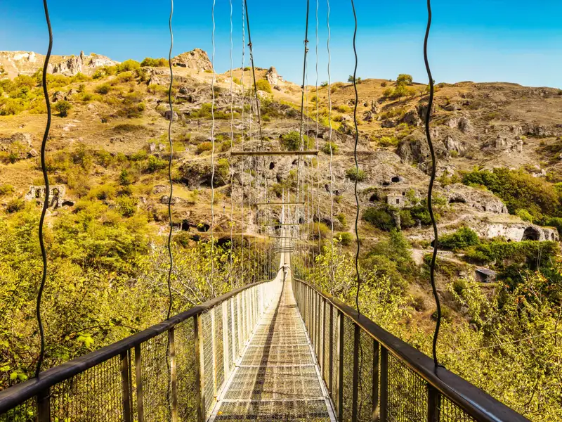 Hängebrücke über eine Schlucht mit historischen Höhlenwohnungen im Hintergrund.