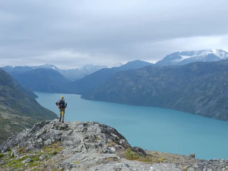 Wanderer auf einem Felsvorsprung mit Blick auf einen türkisfarbenen See und Berglandschaft.
