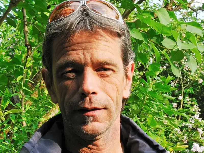 Ein Mann mit Sonnenbrille steht vor grünem Laub.