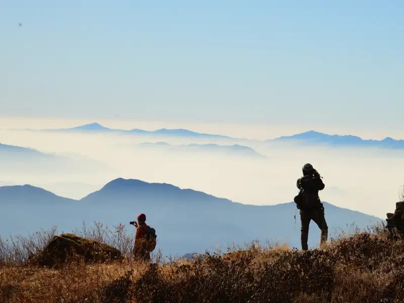 Zwei Wanderer mit Rucksäcken auf einem Berggipfel, die die Aussicht auf die nebligen Berge fotografieren.