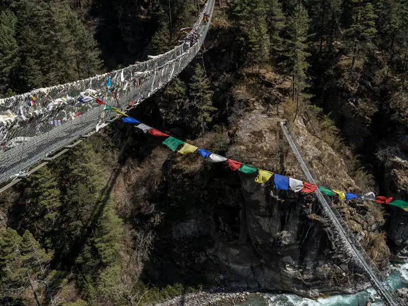 Hängebrücke mit Gebetsfahnen über einer Schlucht.