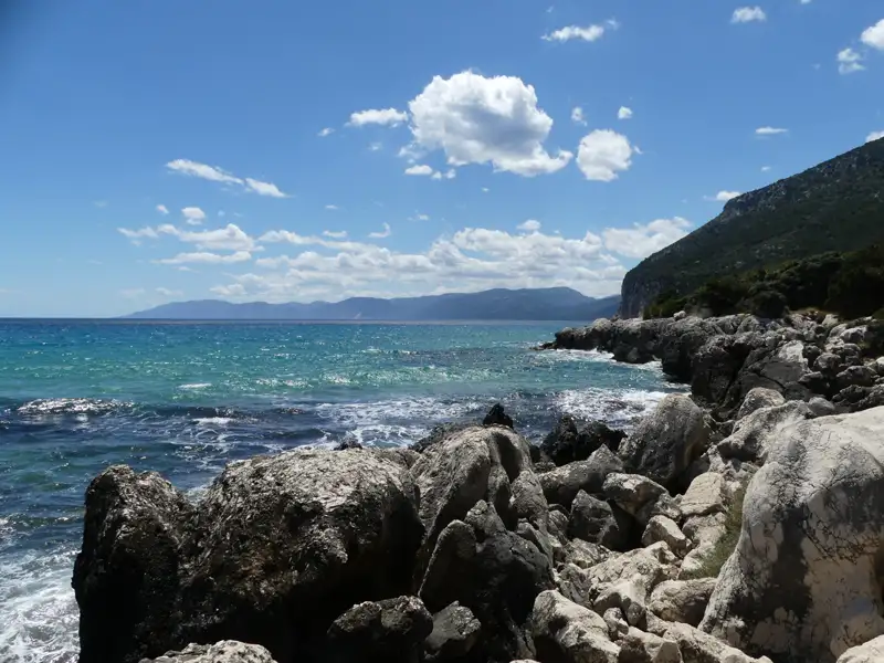 Felsige Küste mit türkisfarbenem Meer und Bergen im Hintergrund.