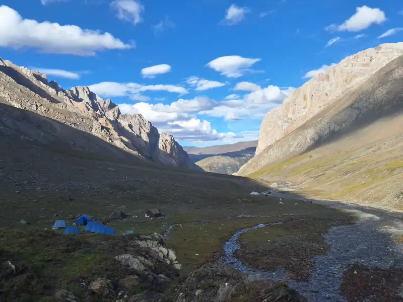 Basislager in einem Gebirgstal, umgeben von hohen Bergen. Blaue Zelte und ein kleiner Fluss sind sichtbar.