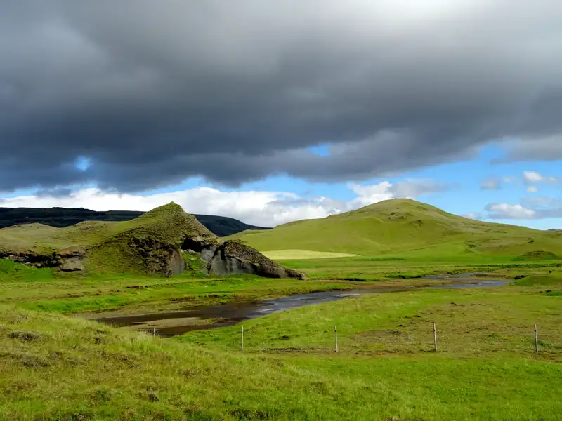 Grüne Hügellandschaft mit Fluss und dramatischem Himmel in Island.