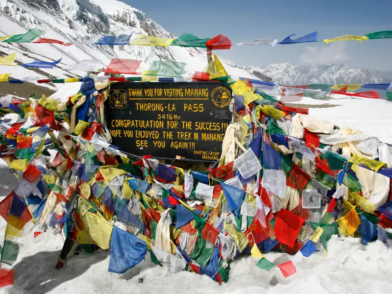 Gebetsfahnen und Schild am Thorong-La Pass (5416 m), der die Grenze zwischen Manang und Mustang markiert und einen Höhepunkt des Annapurna Circuit Treks darstellt.