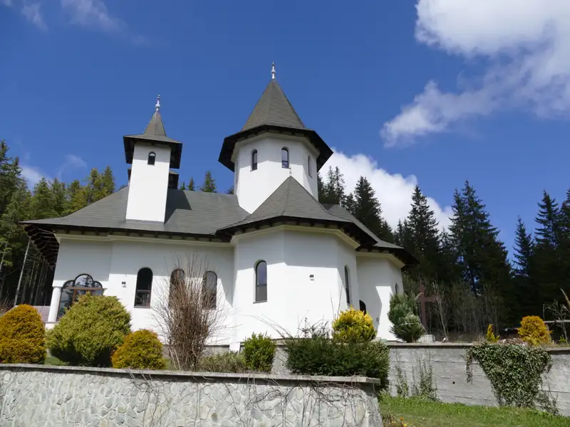 Weiße Kirche mit dunklen Türmen und Dach, umgeben von Nadelbäumen.