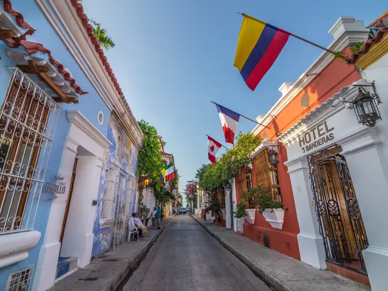 Farbenfrohe Kolonialgebäude säumen eine Straße in Cartagena. Verschiedene Flaggen, darunter die kolumbianische, französische und kanadische, wehen im Wind.