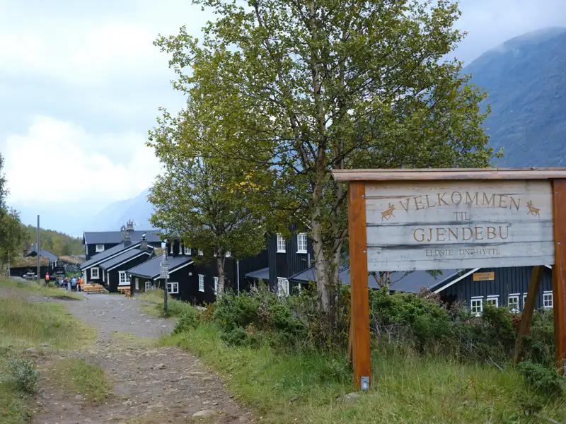 Das Willkommensschild zur Gjendebu DNT-Hütte mit den Hütten im Hintergrund.