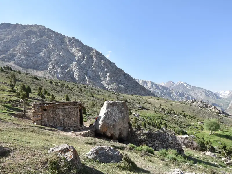 Einfache Berghütte mit Steinmauer und Holzdach vor einer Bergkulisse.