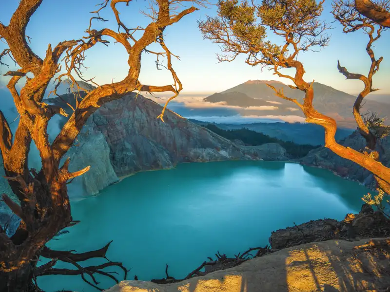 Blick auf einen türkisblauen Kratersee, eingerahmt von Bäumen, mit einem Vulkan im Hintergrund.